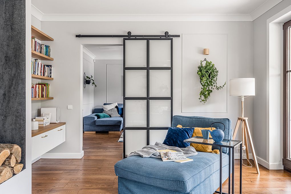 Leseecke, blaues Denim Sofa, Metall- und Glasschiebetüren, Industrietüren, Türöffnung zwischen den Räumen, Holzboden im Wohnzimmer, heller Raum, helle Holzmöbel, blaue und gelbe dekorative Kissen, Wandblumenbeet