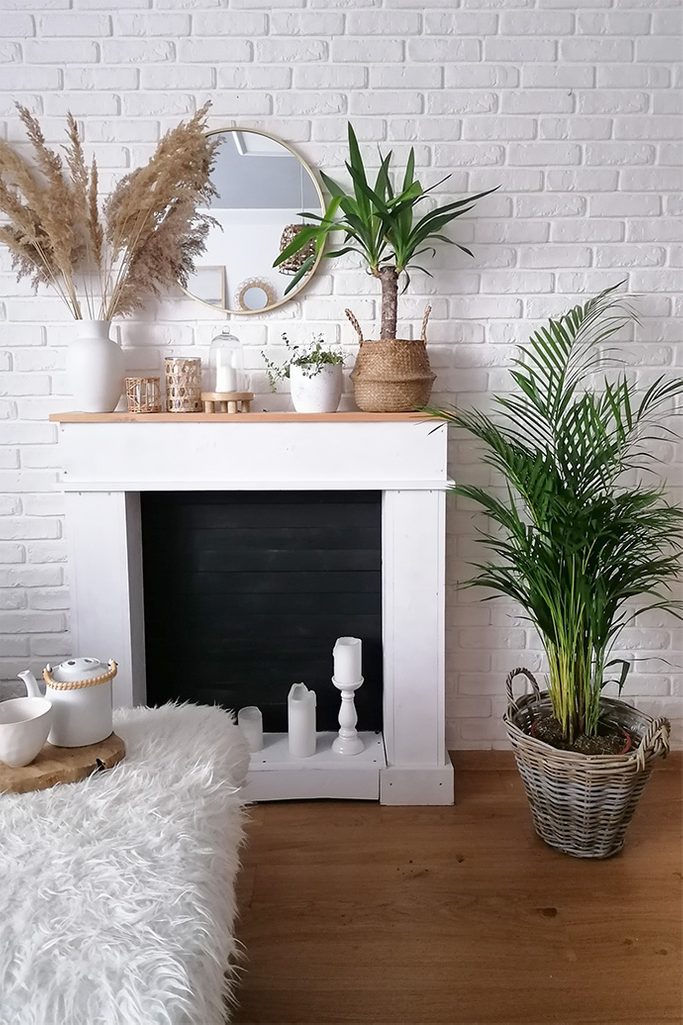 Weiße Ziegelwand, Kamin an einer weißen Wand, Dekorationen am Kamin, Dekorationen im Boho-Stil, große dekorative Pflanze in einem Holzkorb als Raumdekoration