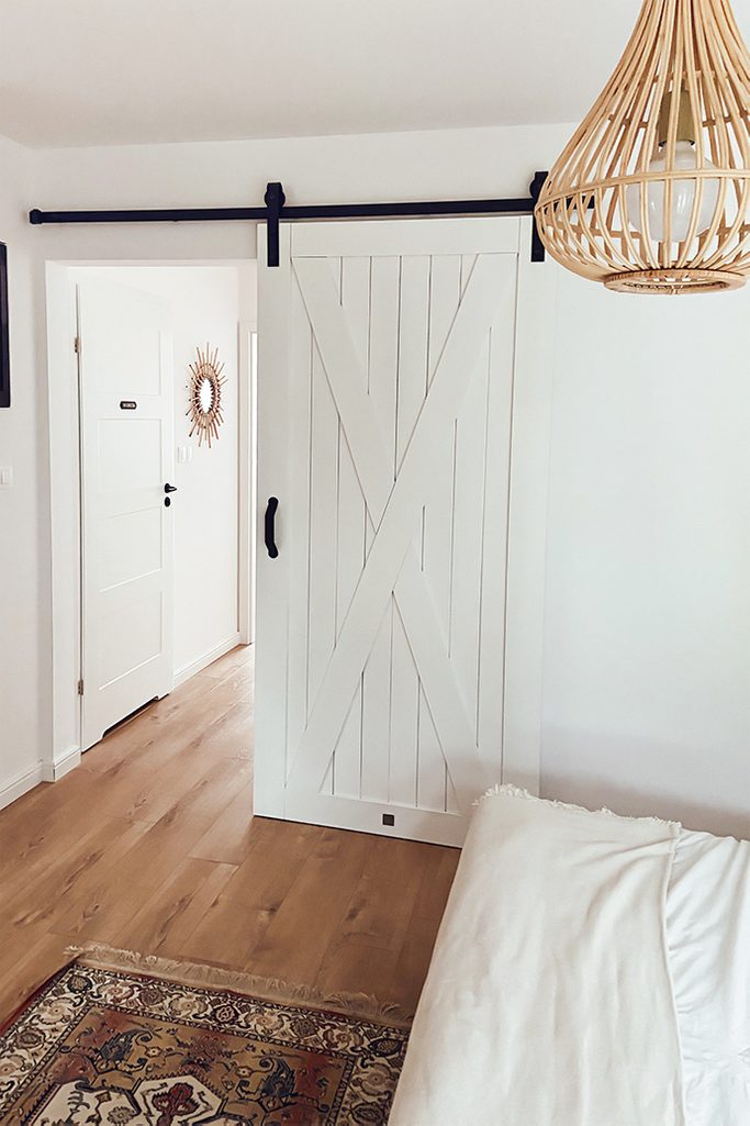 white sliding door in the entrance to the bedroom, white walls and wooden floor in a natural bedroom, bamboo lampshade, colourful, decorative rug in front of the bed