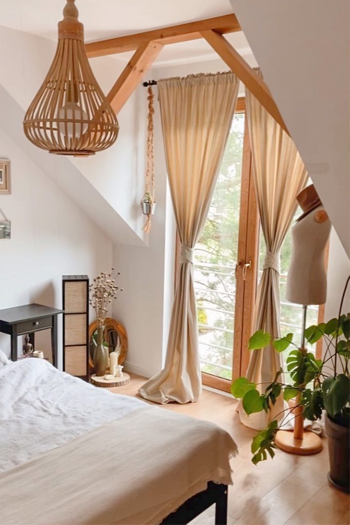 Weiße Wände im Schlafzimmer, beige Vorhänge, Bambus-Hängelampe, schwarzer Nachttisch, Holzbalken an der Decke, Dekorationen im Boho-Stil in einem natürlichen Schlafzimmer