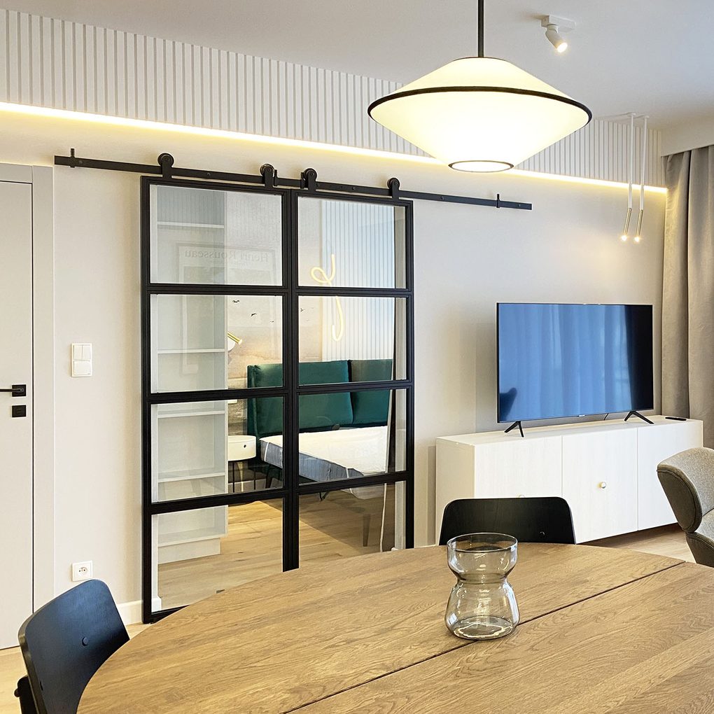 Doppelschiebetüren aus Metall mit Glas, die das Schlafzimmer vom Wohnzimmer in einer modernen Wohnung trennen, Designerbeleuchtung, ein Bett mit gepolstertem Kopfteil, weiße Wände und linearer Stuck