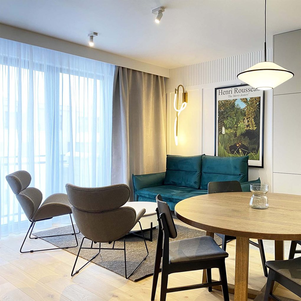 Smaragdgrünes Sofa, graue Sessel auf einem Metallgestell, Wohnzimmer kombiniert mit Essbereich in einer modernen Wohnung, runder Esstisch, moderne Beleuchtung, weiße Wände und graue Accessoires
