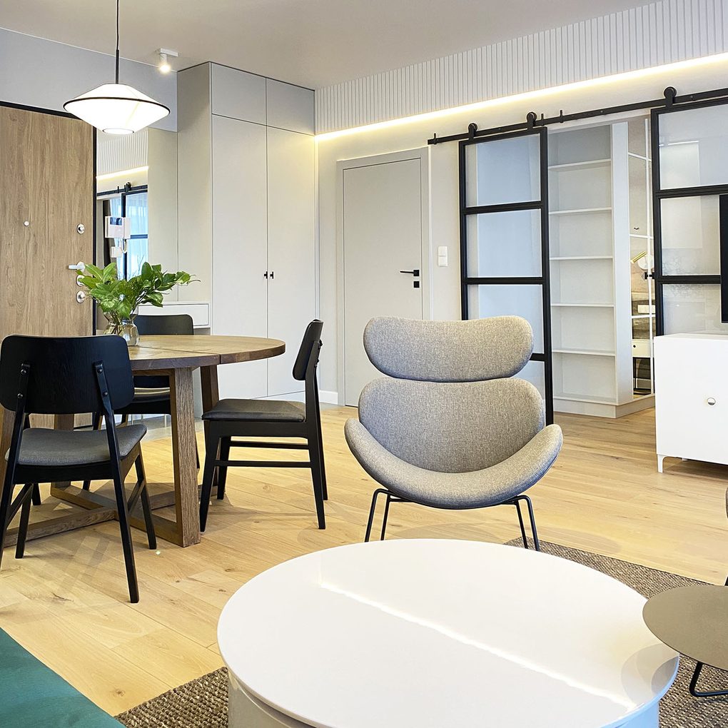 Grauer Sessel auf schwarzem Rahmen, weißer runder Couchtisch, weiße Wände und heller Boden in einer modernen Wohnung, Glasschiebetüren, die das Wohnzimmer vom Schlafzimmer trennen