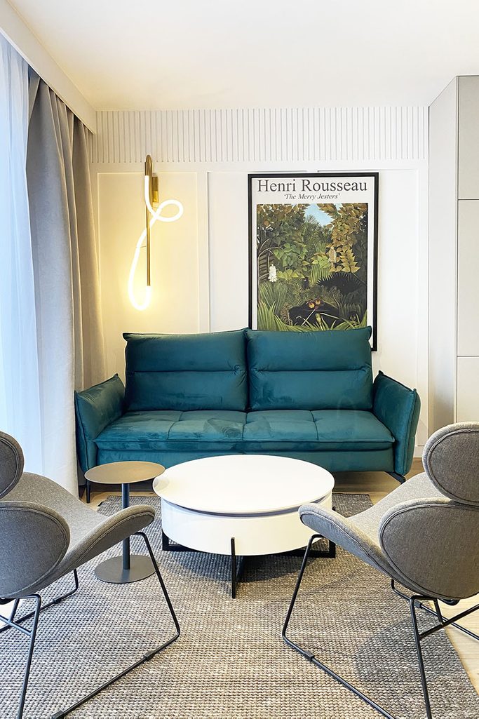 runder weißer Couchtisch, smaragdgrünes Sofa, graue Sessel auf Metallrahmen, moderne Beleuchtung, dekoratives Poster über dem Sofa, linearer Stuck im Wohnzimmer einer modernen Wohnung