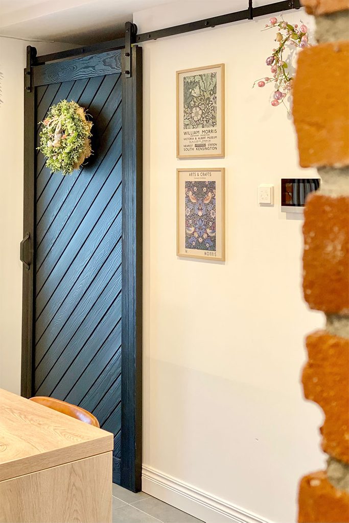 Schiebetür für die Speisekammer, schwarze Schiebetür aus Massivholz, dekorativer Kranz an der Tür, roter Backstein an der Wand, dekorative Gemälde an der weißen Wand