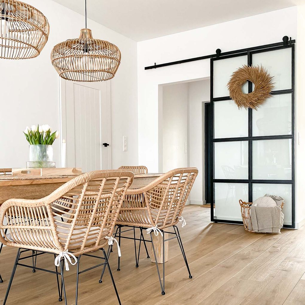 Rattanstühle auf Metallbeinen, ein großer Holztisch, Hängelampen mit geflochtenem Lampenschirm und Glasschiebetüren mit Metallrahmen