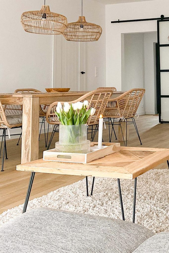 Ein quadratischer Couchtisch auf Metallbeinen und ein großer Holztisch mit Rattanstühlen in einem Wohnzimmer mit weißen Wänden